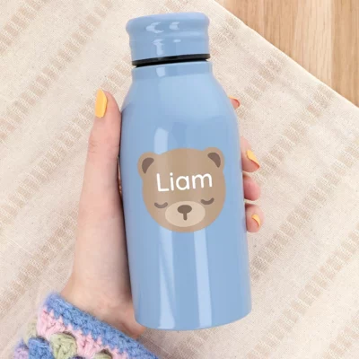 Hand holding a water bottle with a large name label featuring a bear design and the name “Liam”.