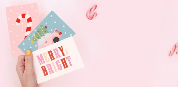 Hand holding colorful birthday cards with holiday designs and the texts “Merry Bright” and “Ho Ho Ho” on a gift.