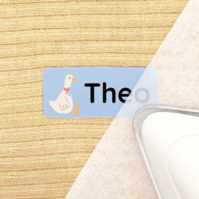 Light blue iron-on label with the name “Theo” and a goose design, placed next to an iron.