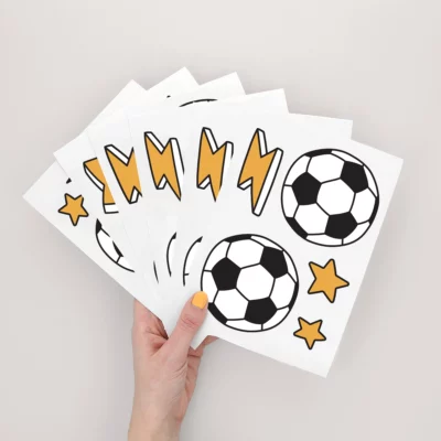 A hand holding colorful soccer-themed stickers featuring black and white soccer balls, yellow lightning bolts, and stars, ideal for children's room decor or personalized name labels.