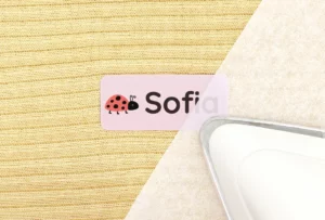 Personalized name tag featuring a ladybug design with the name Sofia, placed on a textured fabric background alongside an iron, ideal for home organization or children's items.