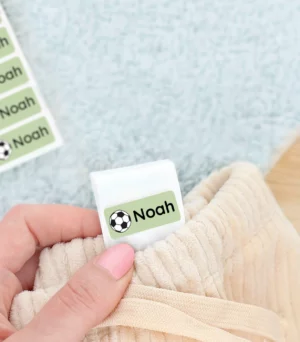 Personalized name tag label featuring a soccer ball design, attached to a child's clothing, with a green design and the name Noah visible. In the background, a pair of green shoes rests on a soft light blue rug.