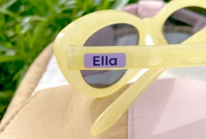Personalized yellow sunglasses with a name tag reading Ella, placed on a soft, colorful background of fabric, surrounded by green grass. Ideal for stylish summer fun and outdoor activities for kids.