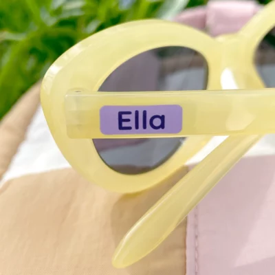 Personalized yellow sunglasses with a name tag reading Ella, placed on a soft, colorful background of fabric, surrounded by green grass. Ideal for stylish summer fun and outdoor activities for kids.