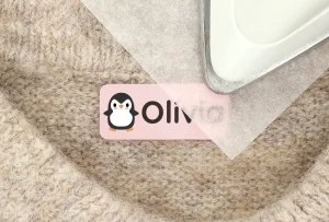 Personalized penguin-themed name tag featuring the name Olivia, placed on a textured fabric surface alongside an iron. Ideal for children's room decor and organization.
