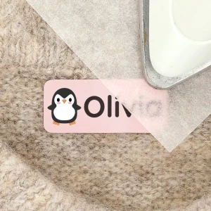 Personalized penguin-themed name tag featuring the name Olivia, placed on a textured fabric surface alongside an iron. Ideal for children's room decor and organization.