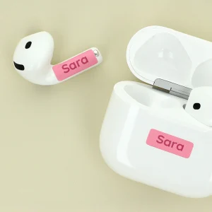Personalized AirPods case and earbud with a pink name tag featuring the name Sara alongside a white charging case on a soft beige background. Perfect for unique gift ideas and personalized accessories.
