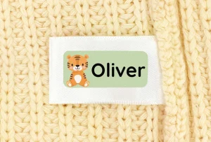 Custom name tag featuring the name Oliver alongside a cute tiger illustration, displayed on a textured beige fabric background. Ideal for personalizing children's belongings.