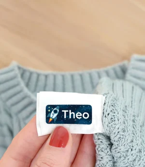 A person holding a personalized name tag with the name Theo, placed beside a cozy sweater, surrounded by colorful autumn leaves on a wooden surface. This image emphasizes custom name tags, perfect for personalizing children's clothing and belongings.